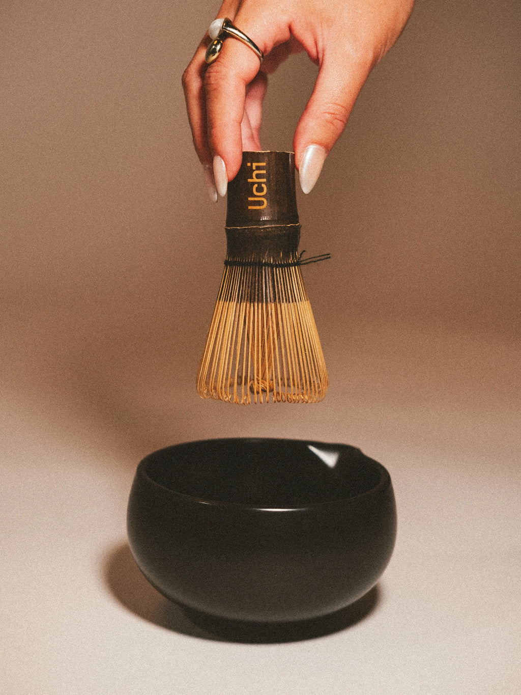 Hand holding a Uchi whisk above a black bowl on a beige background