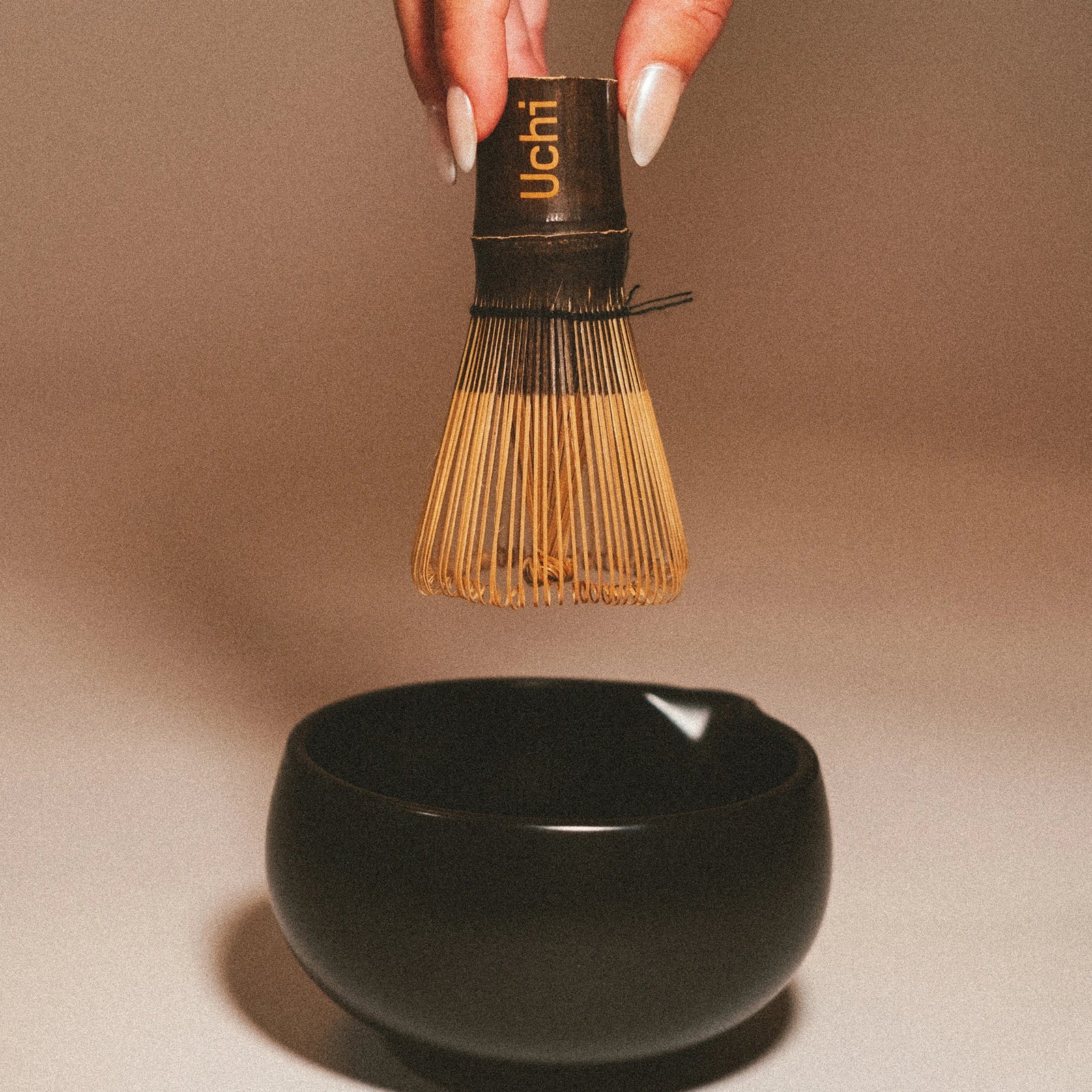 Hand holding a Uchi whisk above a black bowl on a beige background